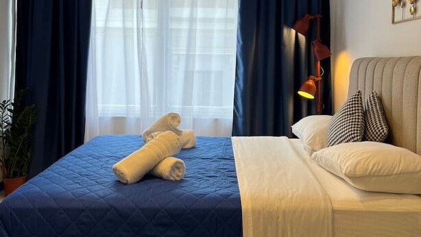 A neatly made bed with blue and white bedding, a bedside lamp, and a view of the outdoors through a window with blue curtains.