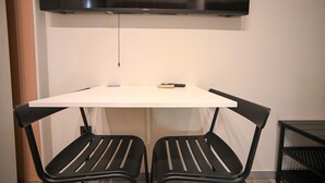 Ein kleines Zimmer mit einem weißen Tisch, zwei schwarzen Stühlen, einem an der Wand befestigten Fernseher und einem kleinen schwarzen Beistelltisch.