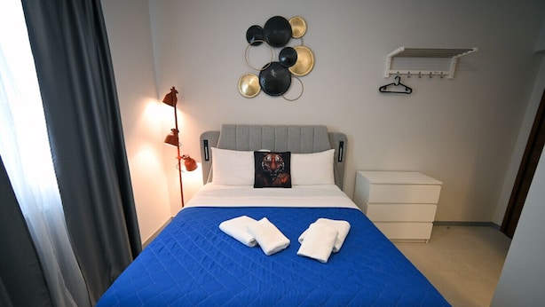 A neatly made bed with a blue bedspread and two white towels, a grey headboard, a wall-mounted light fixture, and a wall decoration of circular plates. - Aura Suite by Zeus suites (Athina)