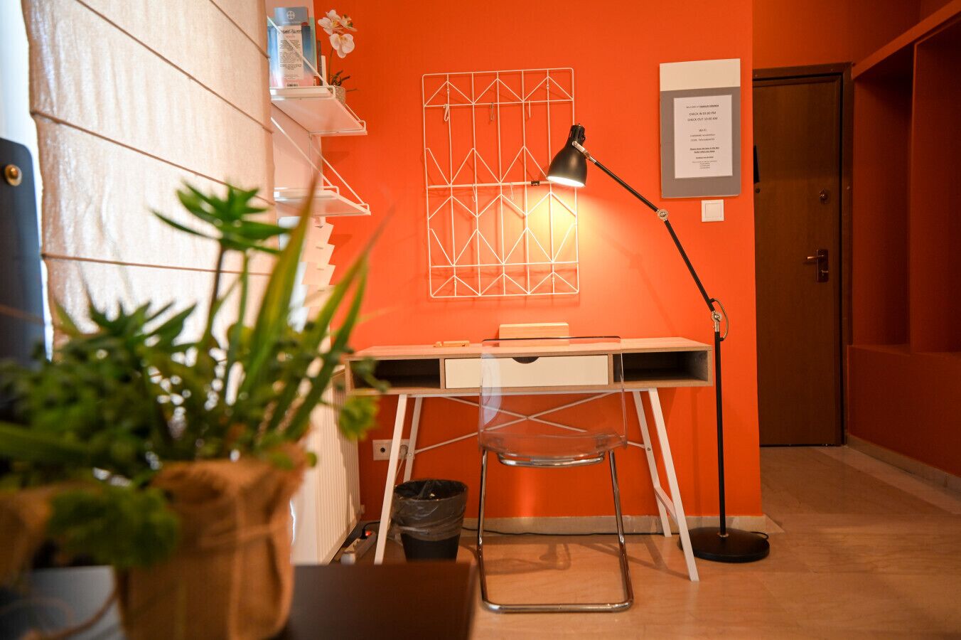 A room with a desk, chair, and a lamp. There is a plant in the foreground and a door in the background.