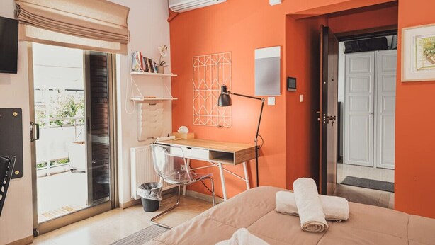 A bedroom with a bed, a desk, a chair, a radiator, a window with a curtain, and a door leading to a balcony.