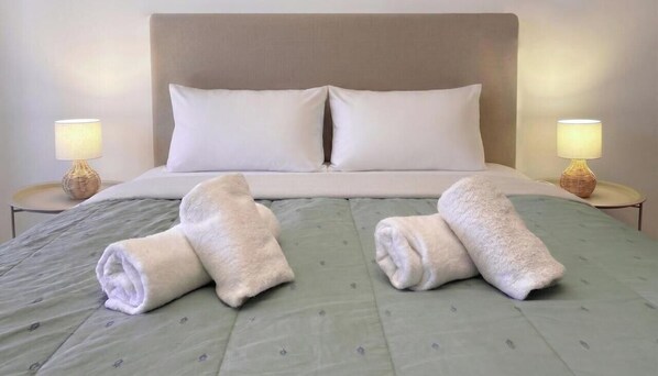 A hotel room with a bed, two pillows, two bedside lamps, and two rolled towels on the bed.