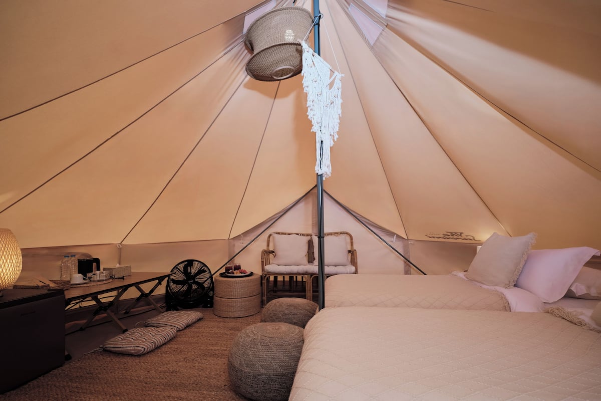 Interior acogedor de una carpa con cama, almohadas, una silla, una mesa con lámpara y un ventilador.