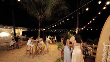 Un área de comedor al aire libre con luces colgantes, palmeras y una barra con botellas.