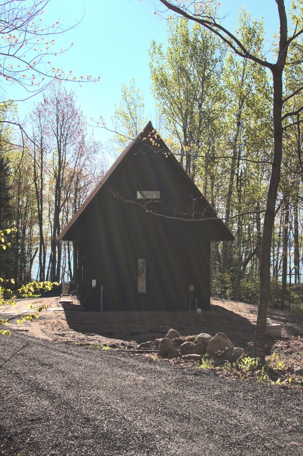A rustic cabin with a sloped roof, surrounded by trees and a gravel path.