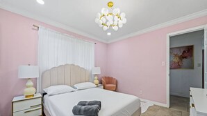 A bedroom with a bed, two bedside tables, a chair, a chandelier, and a door leading to another room.