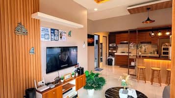Una sala moderna con televisor de pantalla plana, paneles de madera, un comedor con mesa redonda y una cocina al fondo.