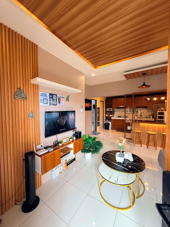 A modern living room with a flat-screen TV, wooden paneling, a dining area with a round table, and a kitchen in the background.