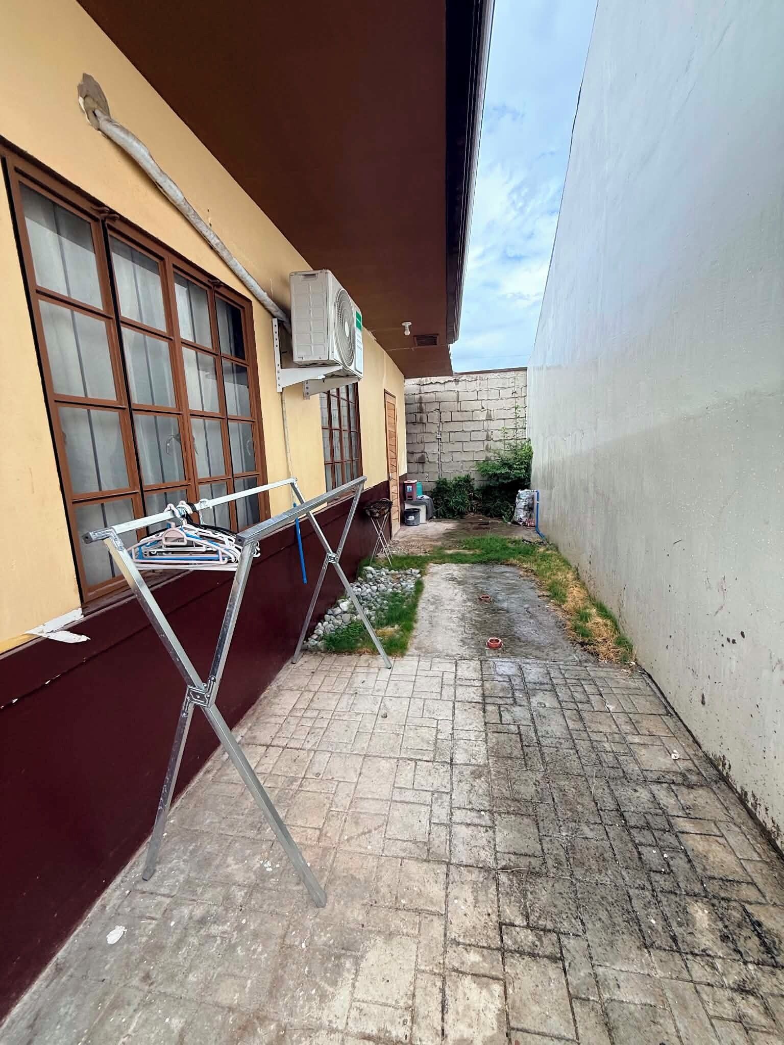 A narrow outdoor corridor with a metal drying rack, a window with blinds, and a brick wall.