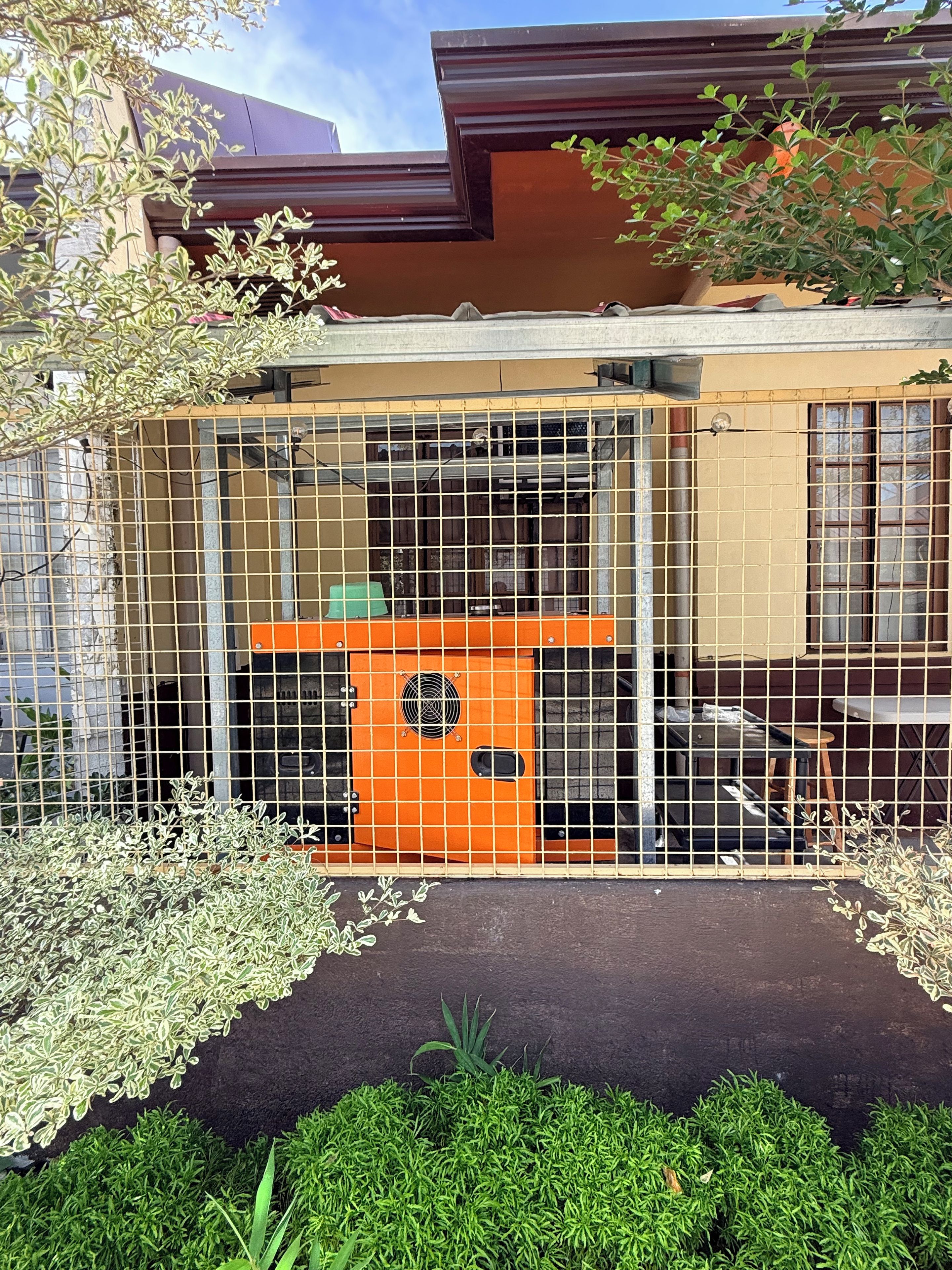 Een omheind buitengebied met een klein oranje gebouwtje, mogelijk een speelruimte of opslagplaats, omringd door groen en een gebouw met een bruin dak.