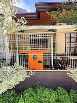 A fenced outdoor area with a small orange structure, possibly a play area or storage, surrounded by greenery and a building with a brown roof.