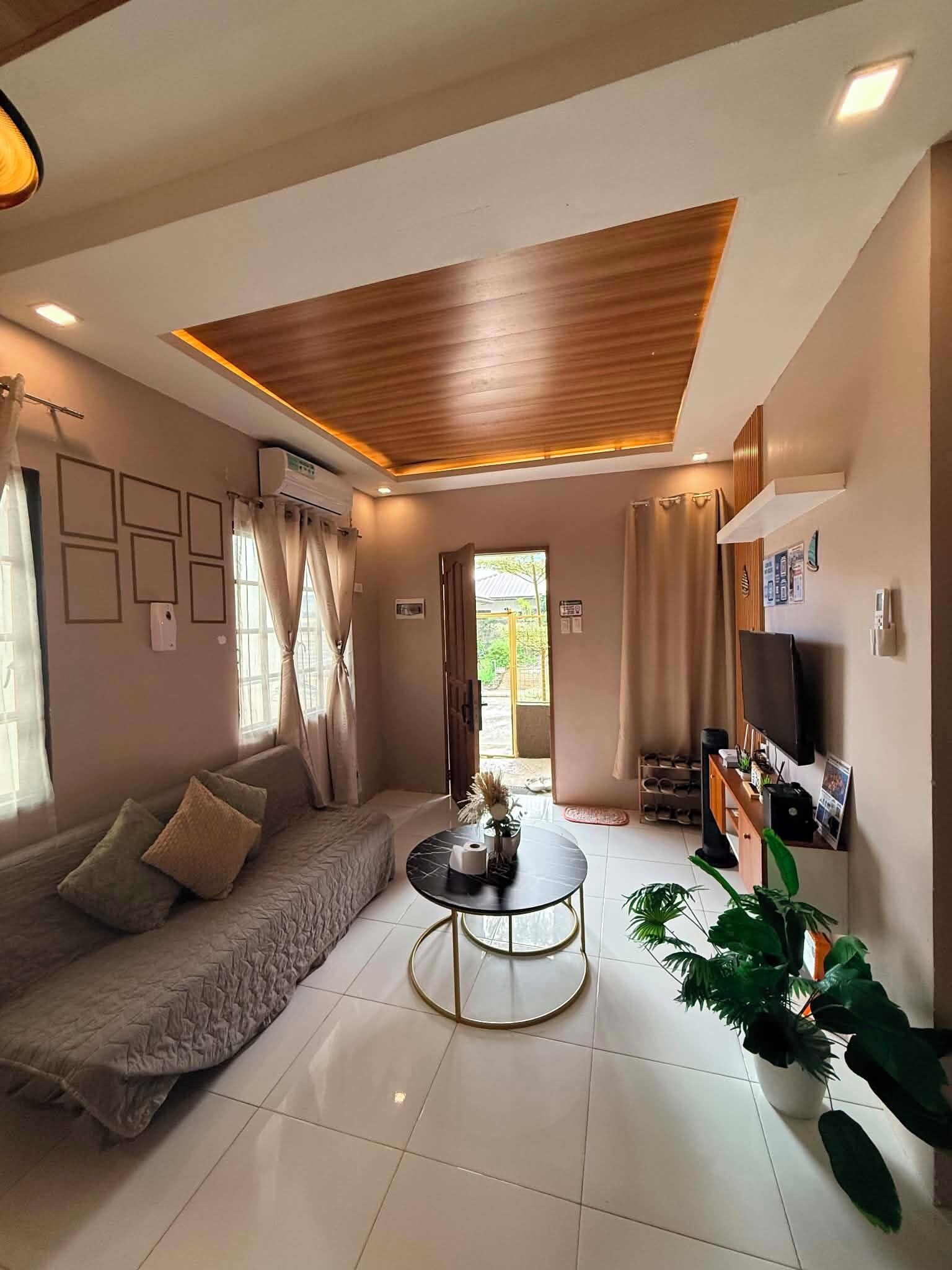 A modern living room with a sofa, a coffee table, and a TV. There is a door leading outside, and the room has a wooden ceiling.
