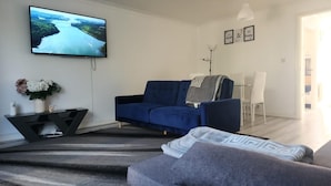 A living room with a blue sofa, a TV mounted on the wall, a small table with a vase of flowers, and a dining area with a white table and chairs. - 3 Bed House - Sleeps 6 - Parking - Wifi (Maidstone)
