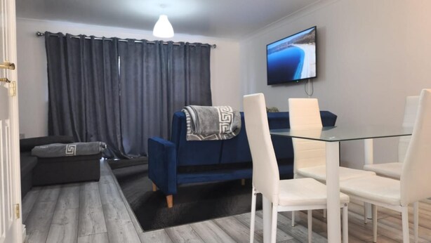 A modern living room with a glass dining table, white chairs, a blue sofa, and a TV mounted on the wall. - 3 Bed House - Sleeps 6 - Parking - Wifi (Maidstone)