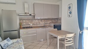 A compact kitchen with a gas stove, white cabinets, and a dining area with a table and chairs. - Apartments with spectacular views of Budoni Bay 3 min. from the sea-4 places (Malamurì)