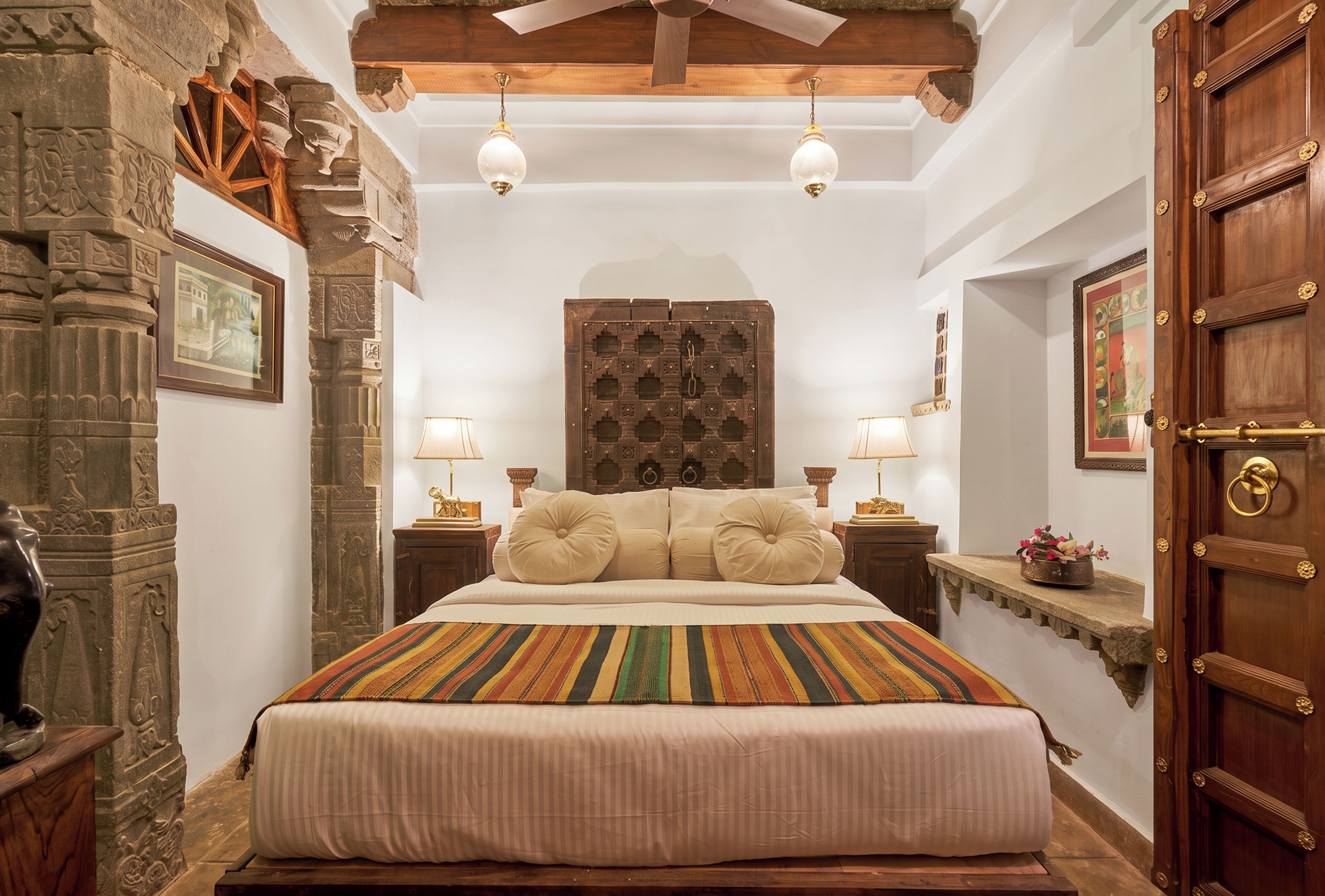 A bedroom with a large bed, two bedside lamps, a wooden door, and a stone wall with carvings.