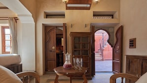 A room with a wooden table, two chairs, a bed, and a view of an outdoor area with a red building. - Kaladwas Lal Haveli (Udaipur)