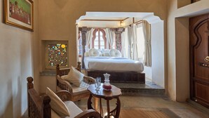 A bedroom with a large bed, a wooden chair, a small table with a vase, and a door. - Kaladwas Lal Haveli (Udaipur)