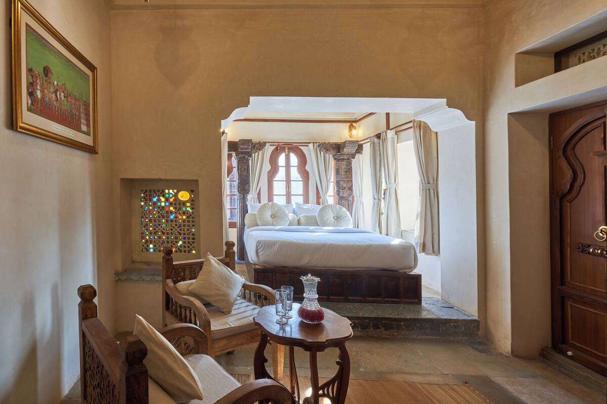 A bedroom with a large bed, a wooden chair, a small table with a vase, and a door.