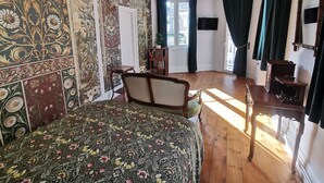 Une chambre avec une couvre-lit à motifs floraux, une chaise en bois, un bureau avec une chaise et une grande fenêtre avec des rideaux.
