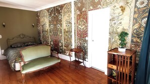 Une pièce avec un plancher en bois, un canapé vert, un lit avec une literie à motifs floraux et un mur recouvert d’un papier peint floral aux motifs complexes.