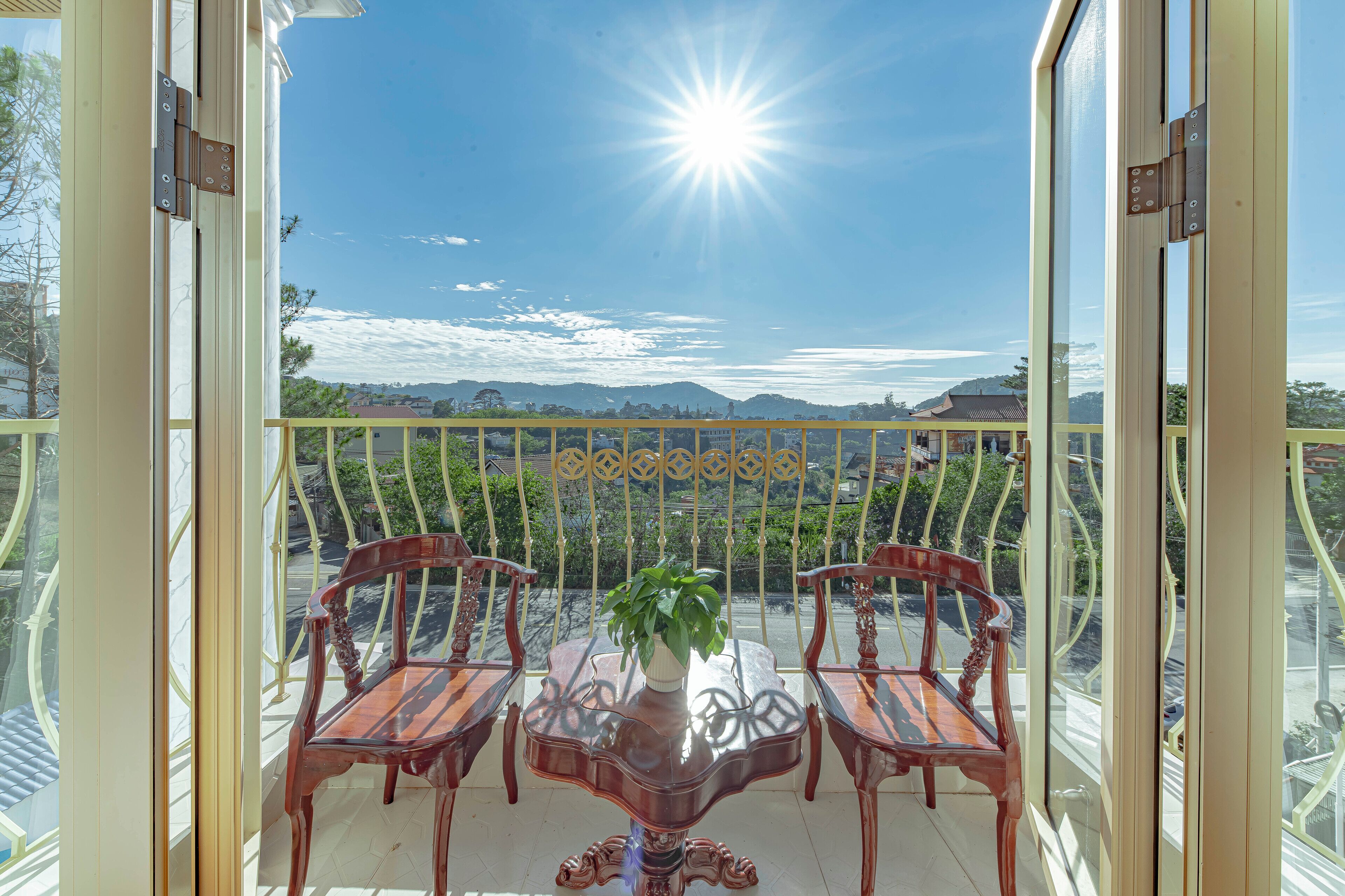 A balcony with two wooden chairs and a table with a potted plant, overlooking a scenic view of a town and mountains.