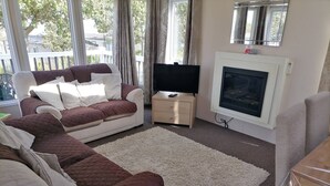 Un salon avec un canapé beige, un canapé blanc, un téléviseur, une cheminée et une vue sur l’extérieur.