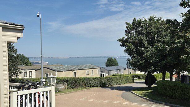 Un quartier résidentiel avec une route asphaltée, un panneau indiquant « Bienvenue au village », et une vue sur la mer en arrière-plan.