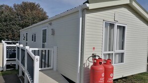 Il s’agit d’une petite maison mobile d’un seul étage, à l’extérieur blanc, avec un petit porche blanc à l’avant et un petit garage attenant. On peut voir deux réservoirs de propane rouges devant la maison.