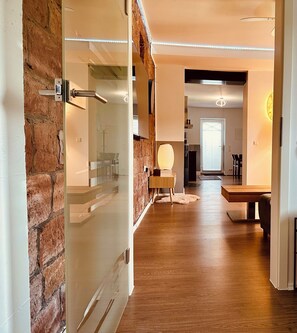 Un umbral abierto que da a una habitación con pared de ladrillo, suelo de madera y un área de comedor con mesa y sillas.