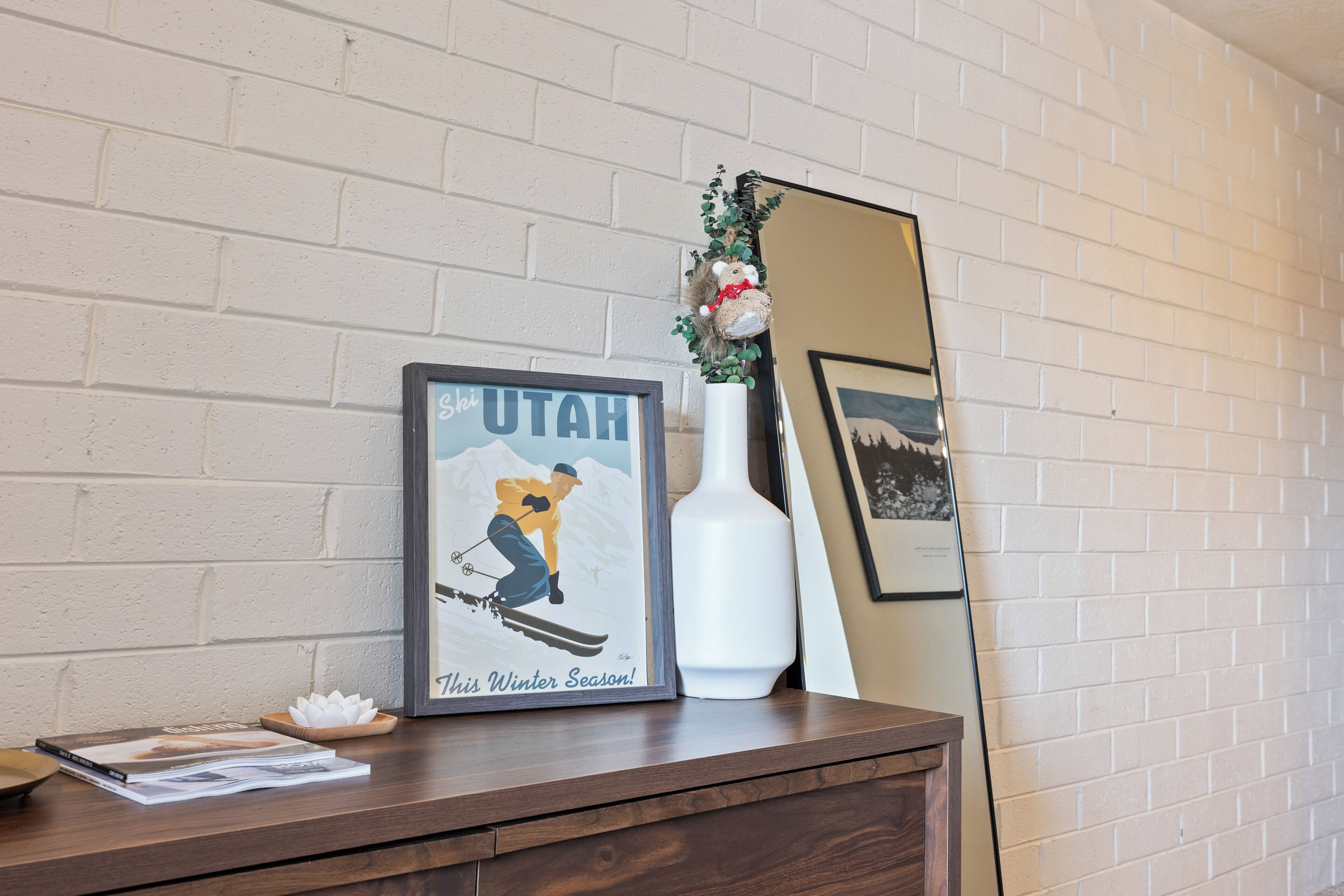A framed poster of a skier on a mountain with the text "This Winter Season!" and a white vase with a plant on a wooden dresser against a white brick wall.