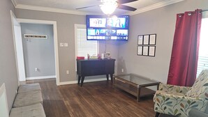 Un salon doté d’un ventilateur de plafond, d’un téléviseur à écran plat fixé au mur, d’une table basse en verre, d’un fauteuil à motifs et d’un parquet en bois foncé.