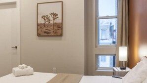 Une chambre d’hôtel avec un lit, une fenêtre donnant sur des immeubles, un tableau représentant des arbres et une table de chevet avec une lampe.