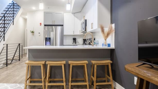 Una cocina moderna con una isla central, taburetes de bar, un televisor y una escalera.