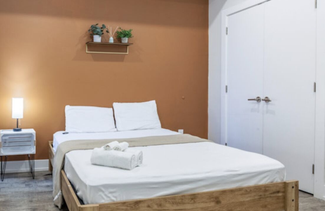 A neatly made bed with white linens and a rolled towel, a bedside table with a lamp, and a shelf with potted plants.
