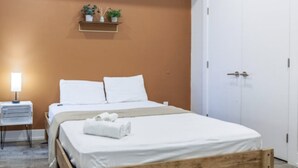 Una cama bien hecha con sábanas blancas y una toalla enrollada, una mesita de noche con una lámpara, y una estantería con plantas en macetas.