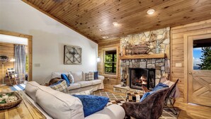 A cozy living room with a stone fireplace, wooden ceiling, and a large window. - Gateway Getaway (Old Fort)
