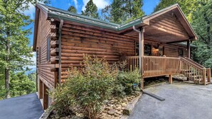A log cabin with a stone foundation, a wooden deck, and a view of the surrounding forest. - Gateway Getaway (Old Fort)