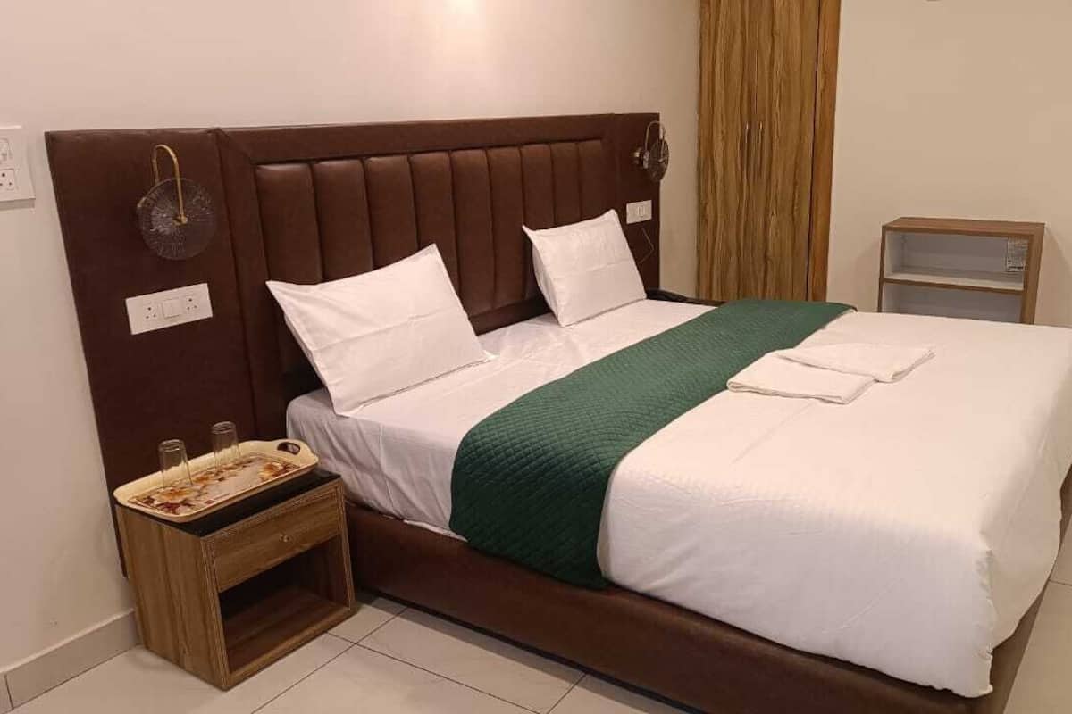 Un lit bien fait, avec une tête de lit marron, des oreillers blancs et une couverture verte. Une table de chevet en bois, sur laquelle se trouve un plateau et une petite étagère, est placée à côté du lit.