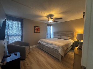 A bedroom with a bed, a chair, a desk, a ceiling fan, and a nightstand with a lamp. - Beach Condo - Less than 75 steps from door to sand! (Cocoa Beach)