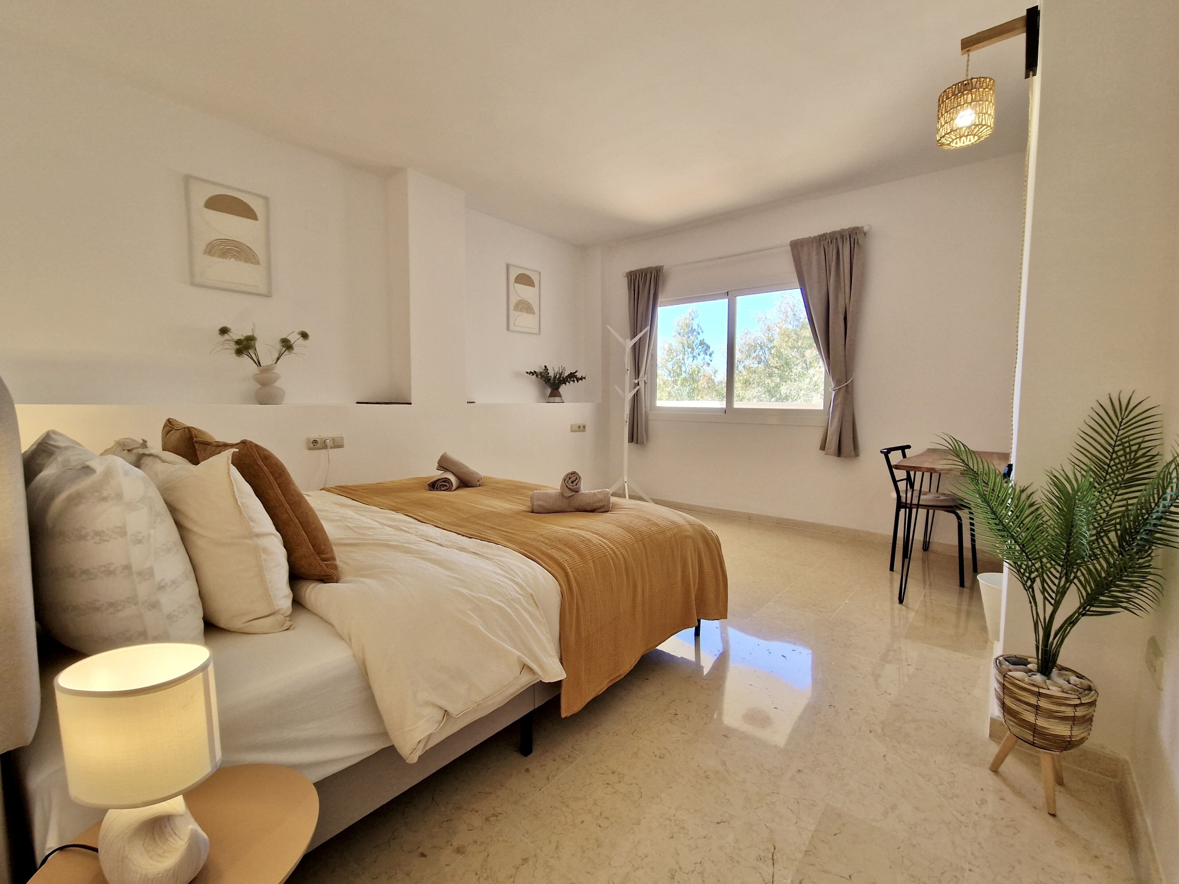 A modern bedroom with a bed, bedside table, lamp, window, curtains, and a small table with chairs.