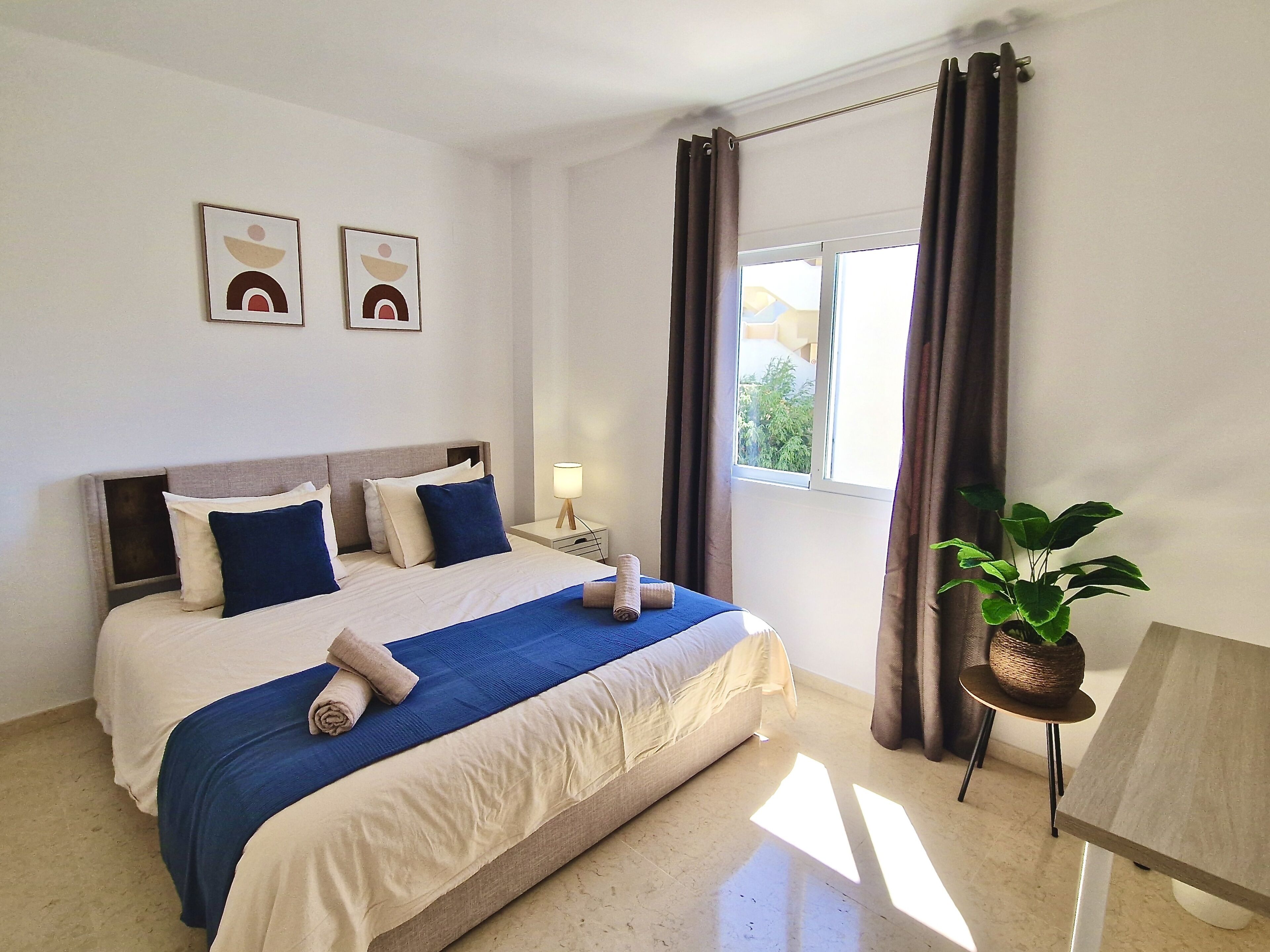 A hotel room with a bed, a nightstand, a lamp, a window with curtains, and a plant.