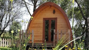 A wooden cabin with a curved roof, a wooden fence, and a garden with yellow flowers. - Glamping Sintra. Enjoy the tranquility of the forest. (Sintra)