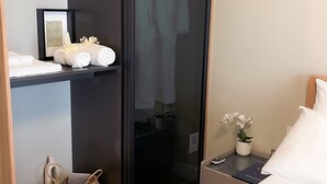 Une chambre d’hôtel comprenant un lit, une table de chevet avec une lampe, une commode, une armoire, une étagère avec des serviettes et un panier, ainsi qu’une plante en pot.
