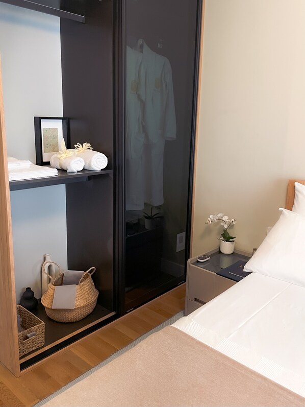A hotel room with a bed, a bedside table with a lamp, a nightstand, a wardrobe, a shelf with towels and a basket, and a potted plant.