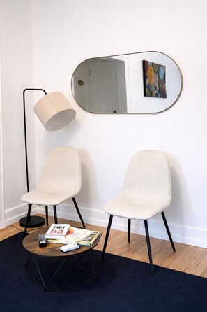 Ein Zimmer mit zwei weißen Stühlen, einem runden Couchtisch mit Büchern, einer Stehlampe und einem Spiegel an der Wand.