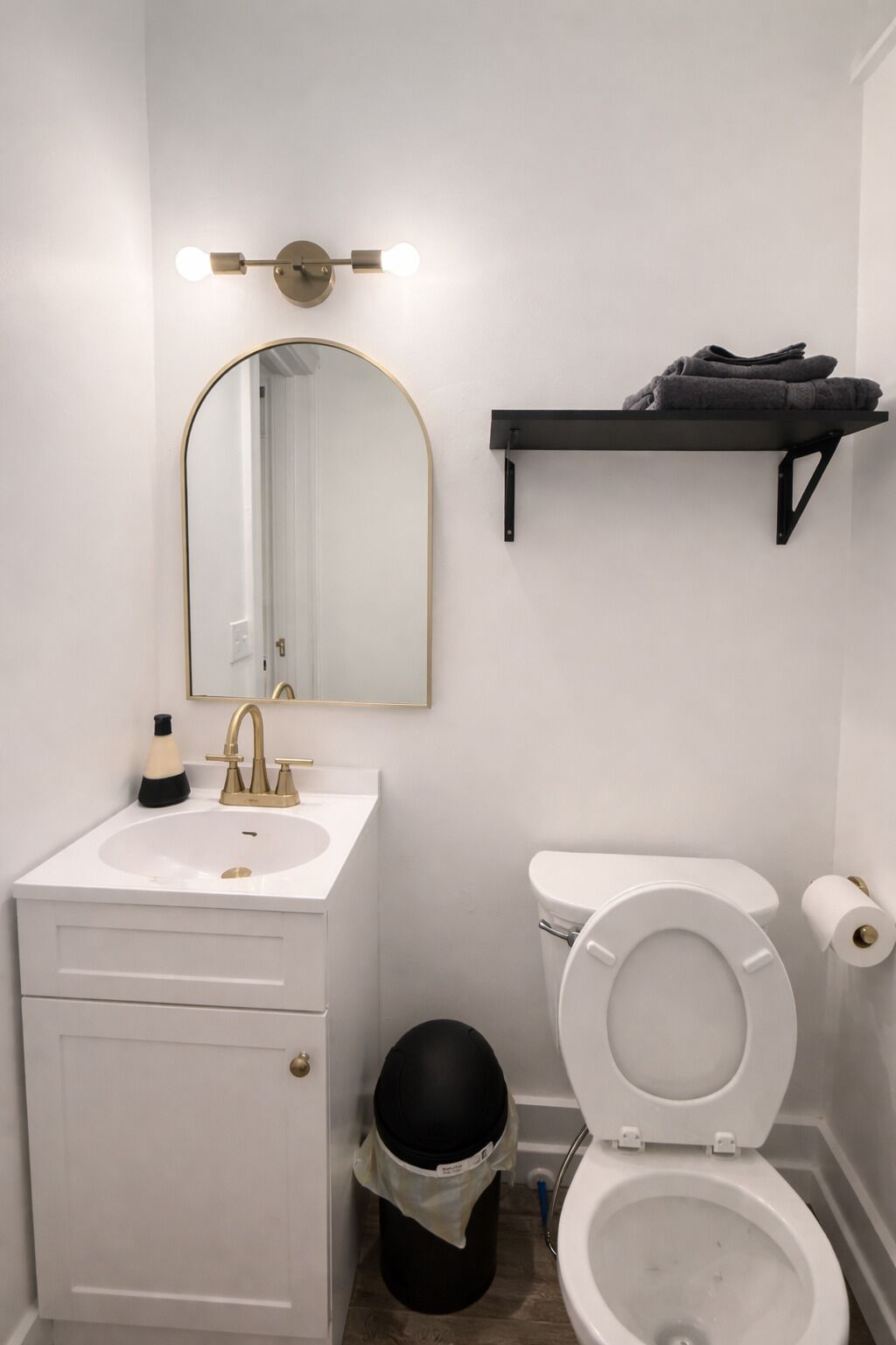 Une salle de bain avec un lavabo blanc, un robinet doré, un miroir avec éclairage intégré, des toilettes, une poubelle et un porte-serviettes.