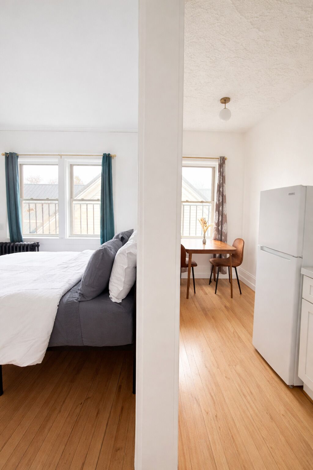 Makuuhuone, jossa on sänky, ruokapöytä tuoleineen, jääkaappi ja ikkuna, jossa on verhot.