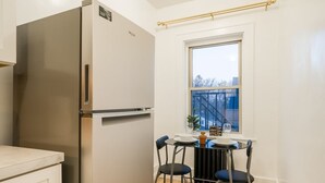 A compact kitchen with a stainless steel refrigerator, a small dining table with two chairs, and a window with a view of buildings outside. - Your Cozy_Studio_HighFalls_TV+Kitchen+Rochest_DT (Rochester)
