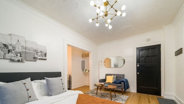 A modern bedroom with a large bed, a sofa, a coffee table, and a cityscape artwork on the wall. - Your Cozy_Studio_HighFalls_TV+Kitchen+Rochest_DT (Rochester)